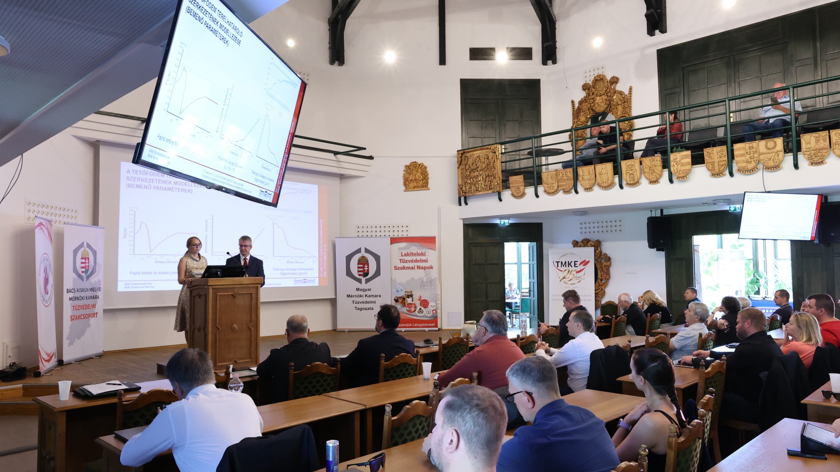 Előadás és közönség a konferenciateremben, a szakmai szervezetek molinóival.